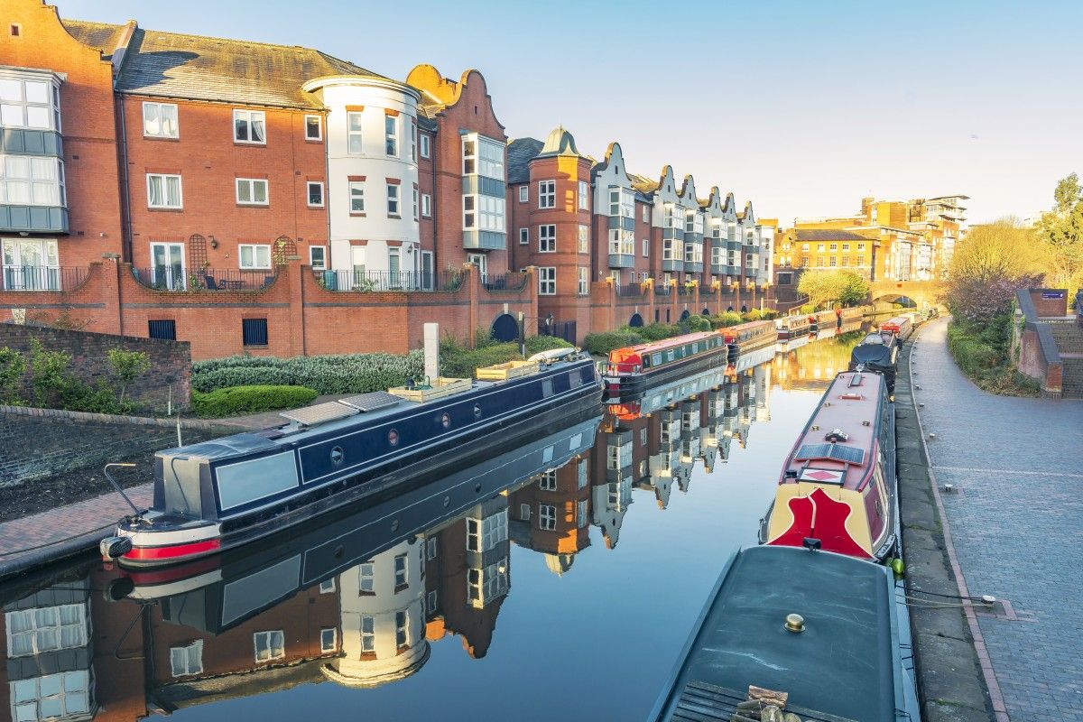 Scenic view of Birmingham canals with colorful narrowboats moored alongside modern brick and white residential buildings reflecting in the calm water.