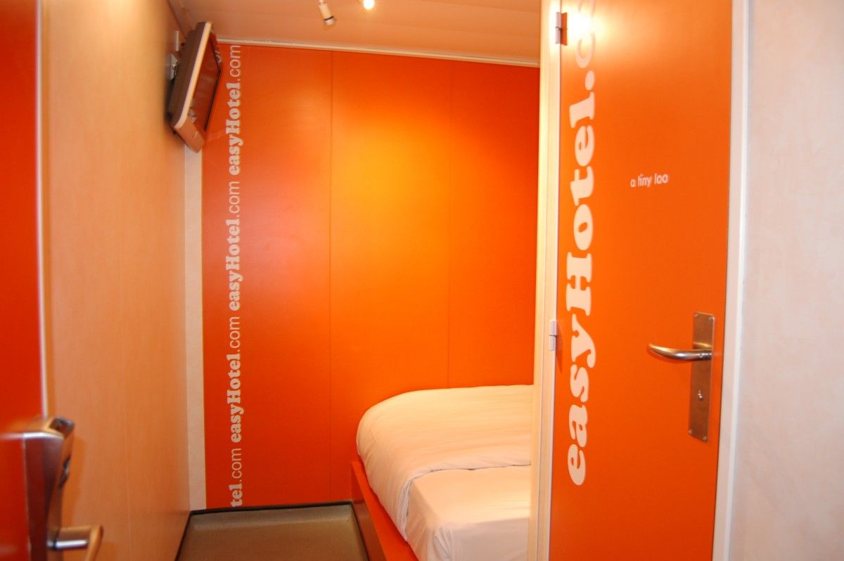 An easyHotel room with bright orange walls, a compact white bed, and a door leading to a small en-suite bathroom.