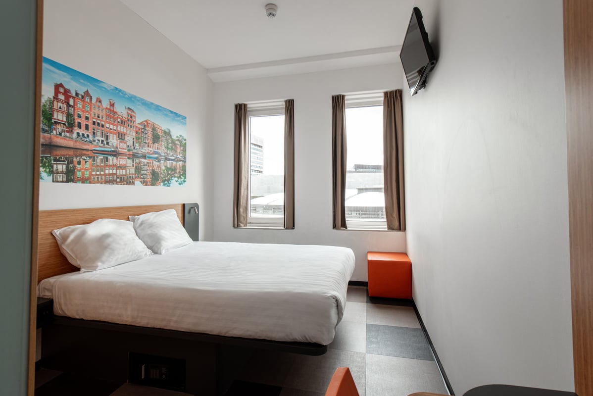 A bright easyHotel Amsterdam Arena double room featuring a large bed with white linens, a wall mural of Amsterdam's iconic canals, two windows with brown curtains, a wall-mounted TV, and an orange cubic stool.