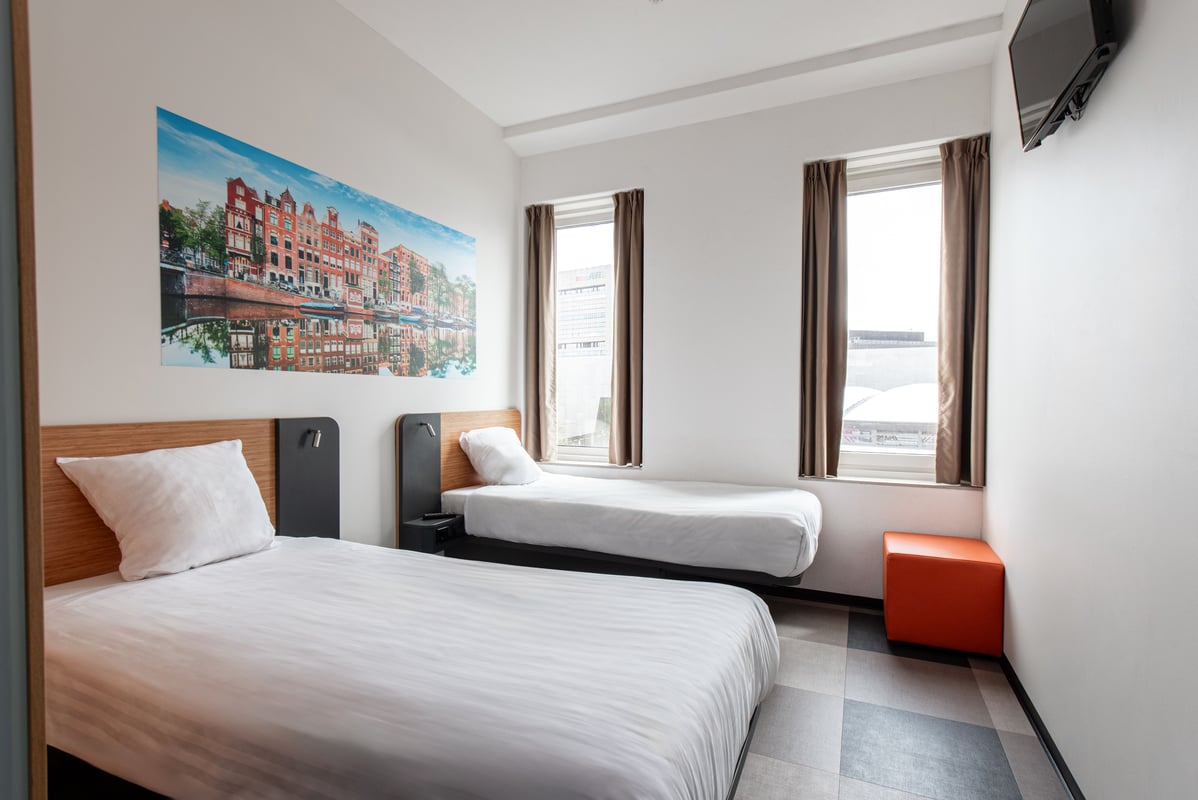 Bright easyHotel Amsterdam Arena twin room featuring two single beds with white linens, a large wall mural depicting Amsterdam canal houses, and a vibrant orange footstool.