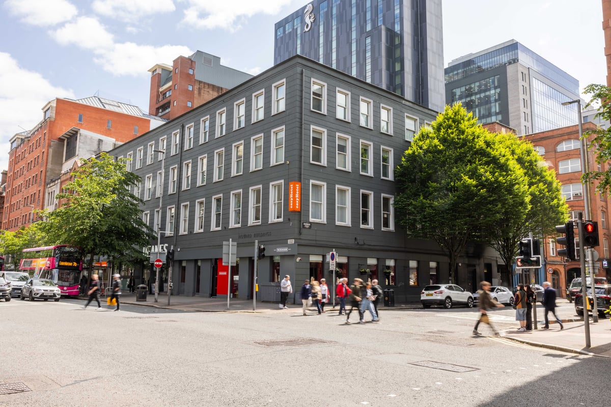 Exterior view of the easyHotel Belfast building on a city street corner. The multi-story hotel is dark grey with many white-framed windows, with pedestrians and a red telephone box visible on the street.