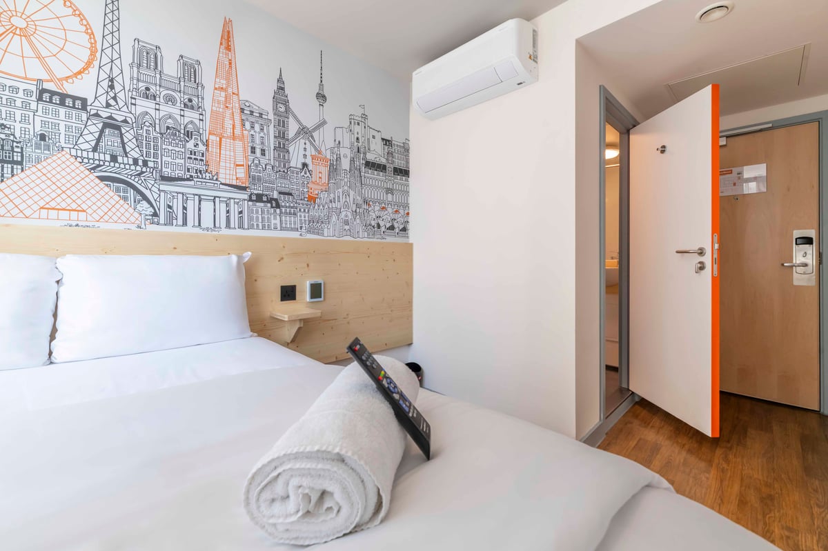 EasyHotel Croydon double room with white linens, pillows, and a rolled white towel on the bed. The wall features a city skyline mural.