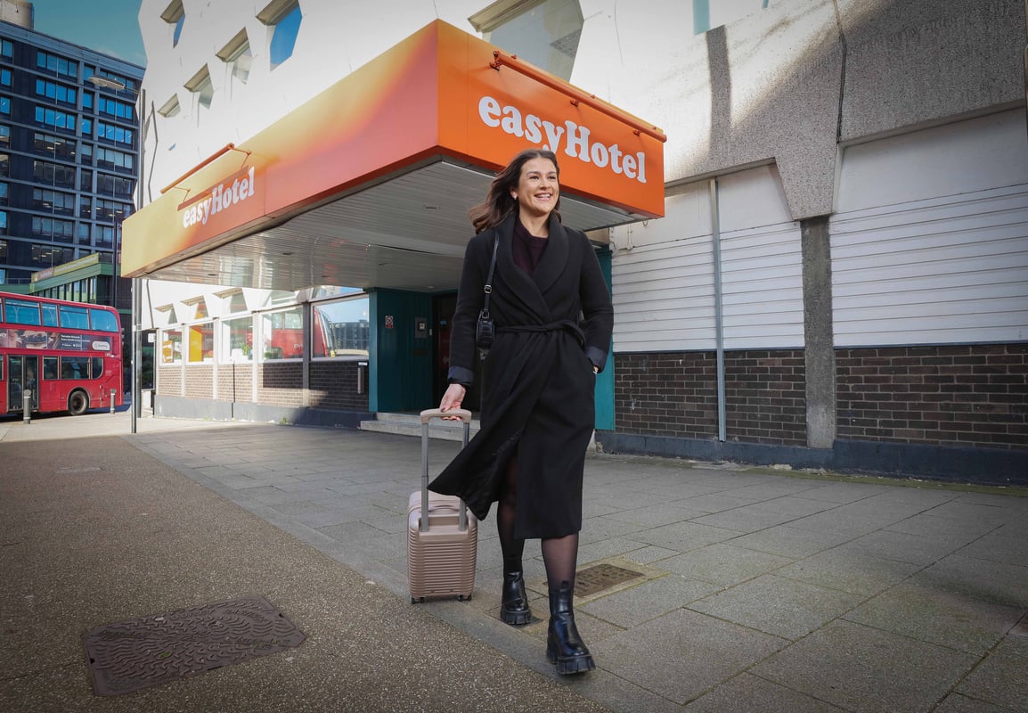 Person exiting a simple hotel entrance in Croydon, London, with a suitcase and a red bus in the background.