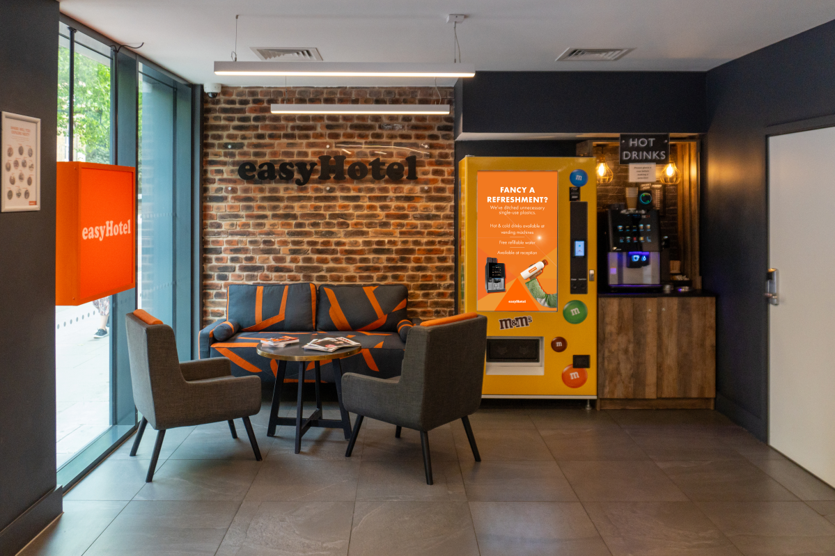 easyHotel London City Shoreditch reception and lobby, featuring a comfortable lounge area with grey and orange seating, an exposed brick wall with the hotel logo, large windows, and a dedicated refreshment station with hot drinks and snack vending machine