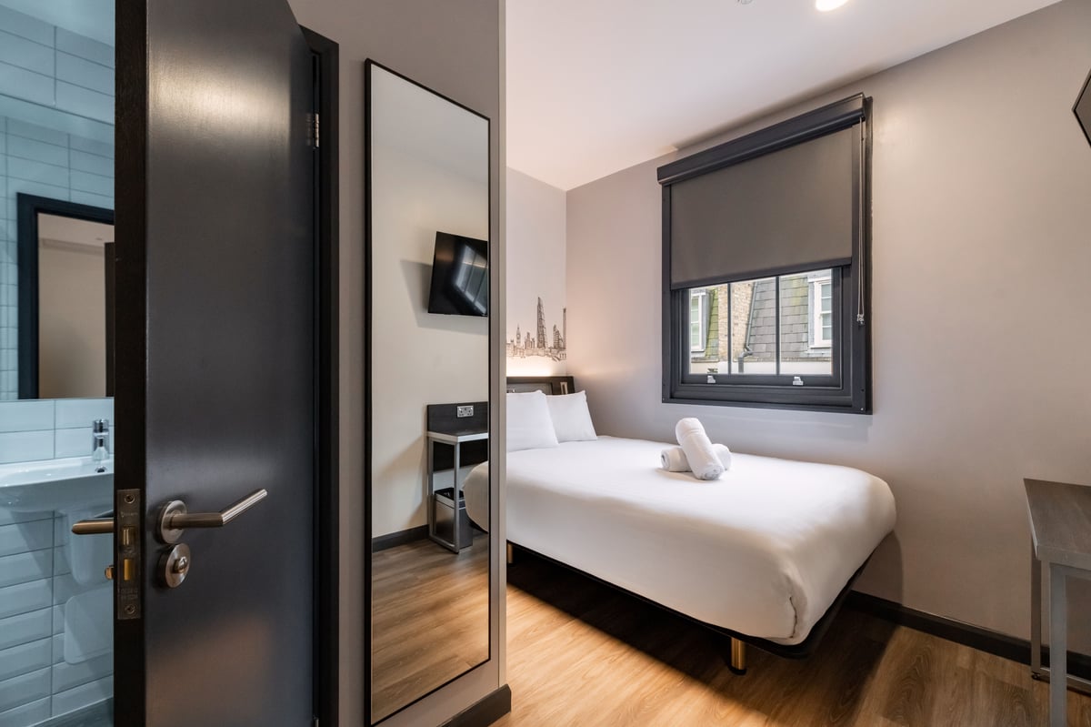 A modern, compact easyHotel family room in London Victoria featuring a bed with white linens and towels, a large window with a dark blind, a wall-mounted TV, and an adjacent bathroom.