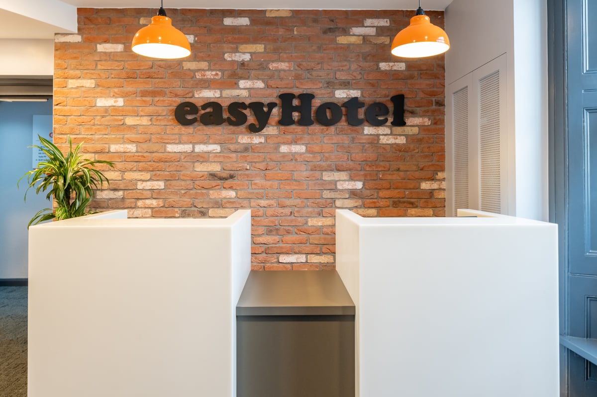 A modern easyHotel London Victoria reception area featuring a white desk, an exposed brick wall with the easyHotel logo, and bright orange pendant lights.
