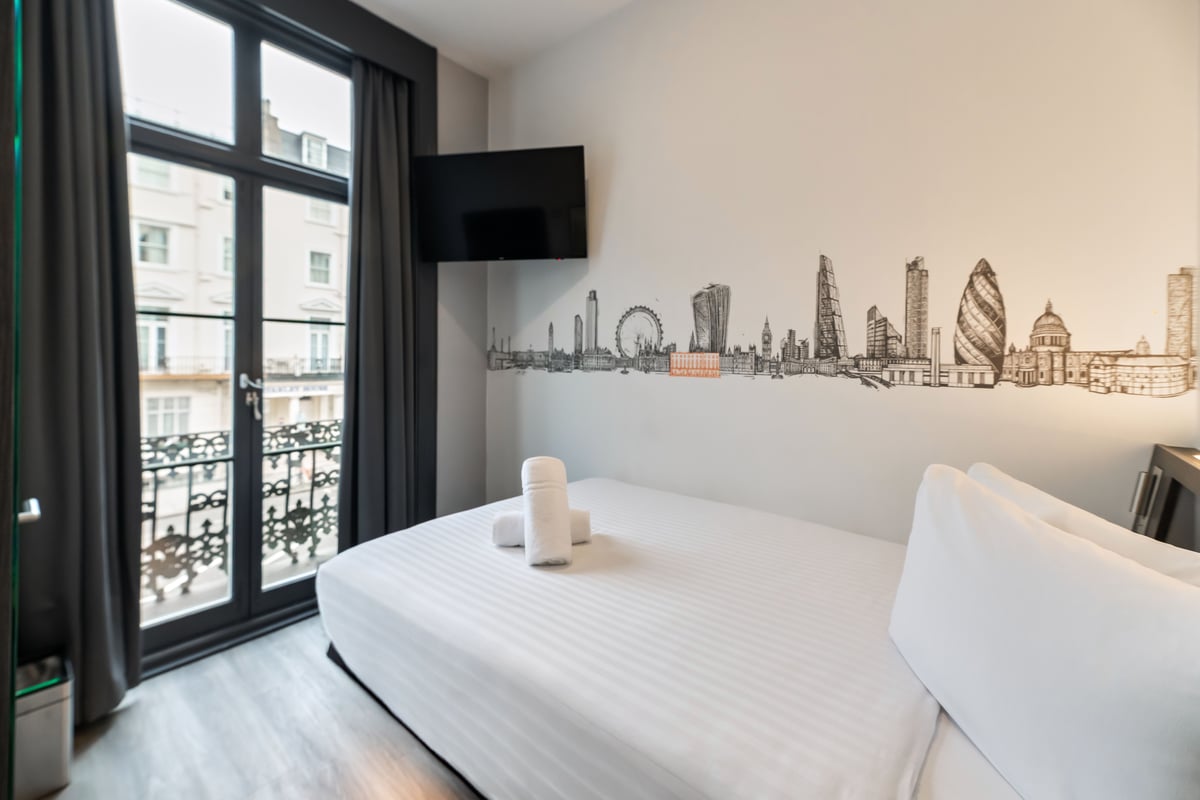 easyHotel London Victoria Standard Double Room with a white double bed, iconic London skyline wall art, a mounted flat-screen TV, and large windows opening to a decorative balcony.