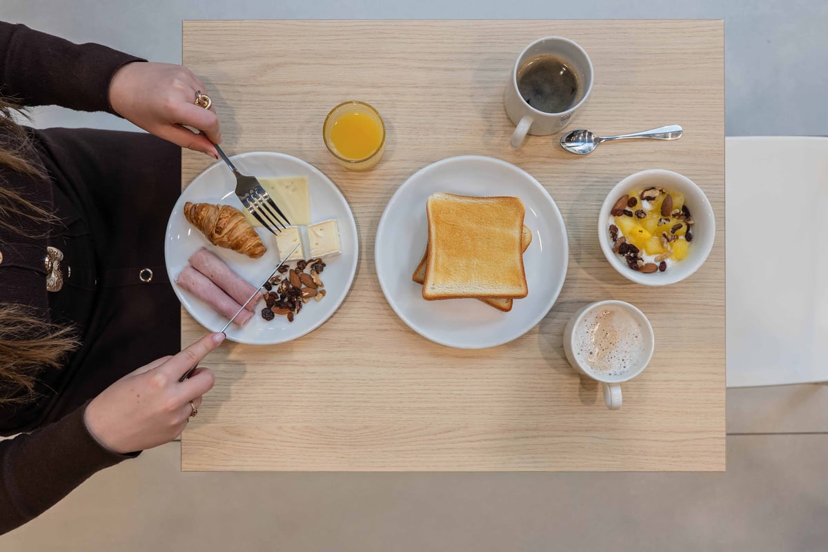 EasyHotel Spanish & French Breakfast: A top-down view of a simple breakfast on a light wood table, featuring toast, sausage, croissant, cheese, fruit salad, orange juice, and coffee.