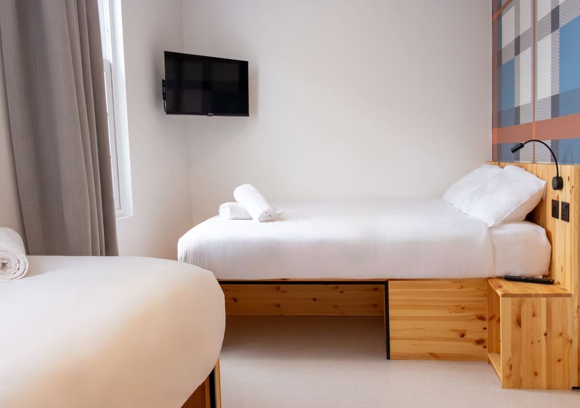 A bright easyHotel Paddington triple room featuring two single beds with white linens, a wall-mounted TV, and a distinctive blue and orange plaid accent wall.