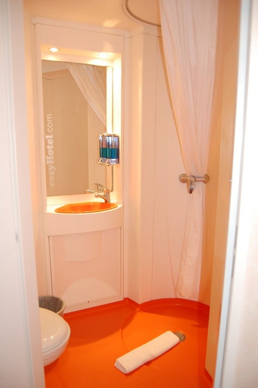 A compact easyHotel bathroom featuring a distinctive orange floor, integrated sink with an orange basin, toilet, mirror, and a shower area with a white curtain.