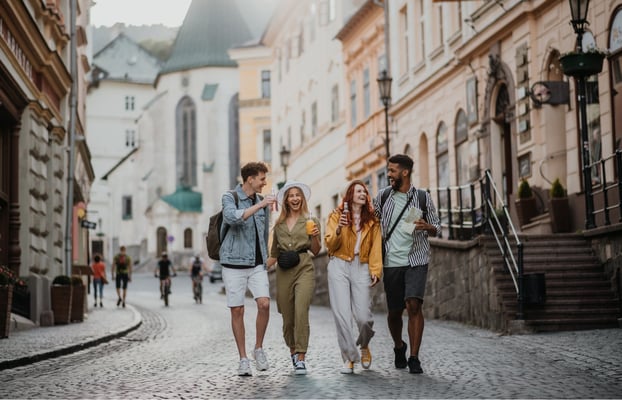 Four smiling young adult friends exploring a historic cobblestone street in Banská Štiavnica, Slovakia, with traditional architecture and a prominent church building.