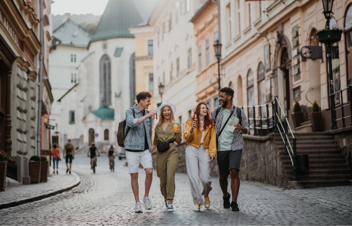 Four diverse young friends laughing and walking on a cobblestone street in a picturesque historic European city, surrounded by classic architecture and a prominent church, perfect for group travel and tourism marketing.