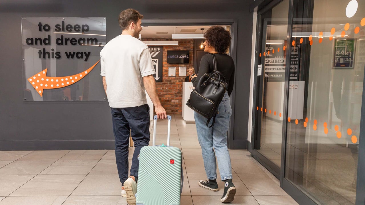 Two travelers with a suitcase and backpack entering a modern hotel reception area. A wall sign with an orange arrow reads 'to sleep and dream, this way'.
