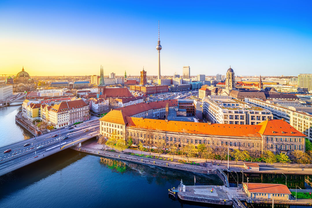 Panoramic aerial view of Berlin, Germany, at sunrise, showcasing the Spree River, bridges, historic buildings with terracotta roofs, and the distinctive Berlin TV Tower in the background.