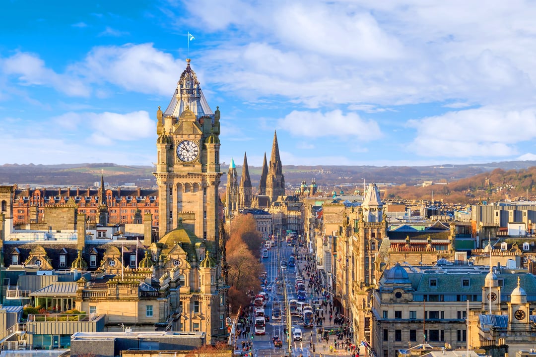 Panoramic cityscape of Edinburgh, Scotland, looking down Princes Street with its iconic clock tower, historic spires, and busy street filled with people and vehicles under a bright blue sky.