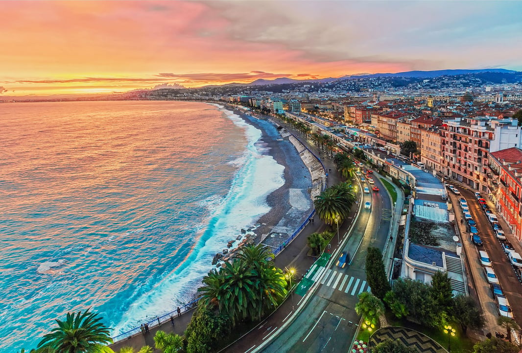 Panoramic view of Nice, France, at sunset, showcasing the vibrant Mediterranean coastline, Promenade des Anglais with palm trees and traffic, and the city buildings extending towards the mountains.