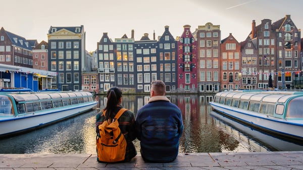 Two people sit on a canal-side walkway in Amsterdam, Netherlands, overlooking historic canal houses at sunset. Canal boats are docked in the canal.